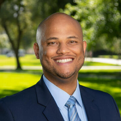 Smiling man in a dark suit and blue tie standing outdoors with trees and grass in the background.