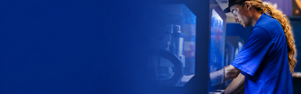 Student operates advanced manufacturing equipment in a lab, with a blue gradient overlay fading from left to right and creating open space for webpage content. The background features industrial machinery and a collaborative engineering workspace.